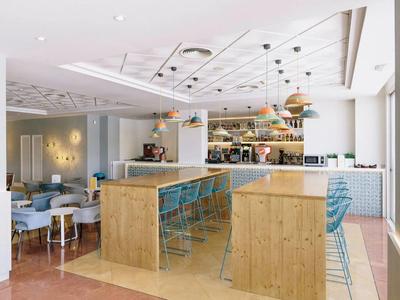 Area bar moderna dell'hotel con banconi in legno, sgabelli blu e zona soggiorno accogliente sullo sfondo.