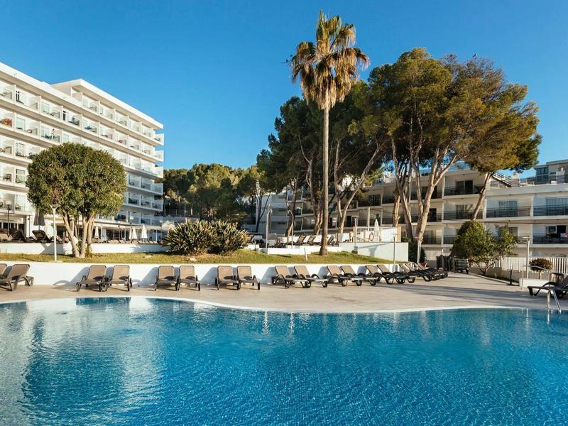 Hotel con grande piscina, lettini prendisole e palme sotto un cielo azzurro limpido.