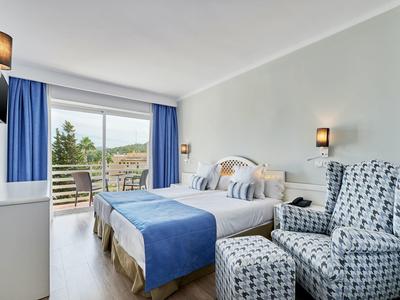 Camera d'hotel luminosa con letto matrimoniale, copriletto blu, poltrone e balcone con vista sul verde.