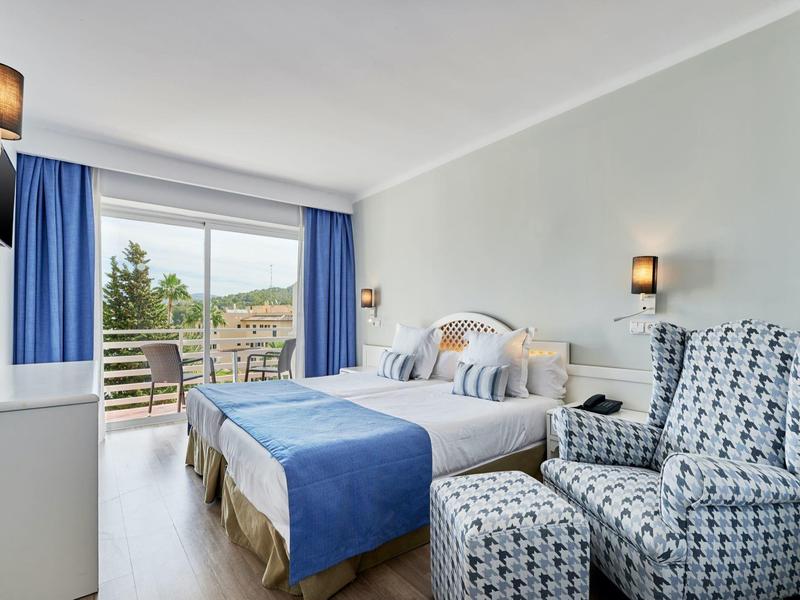 Camera d'hotel luminosa con letto matrimoniale, copriletto blu, poltrone e balcone con vista sul verde.