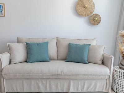 Canapé beige avec deux coussins bleus devant un mur blanc avec décoration murale et vase.