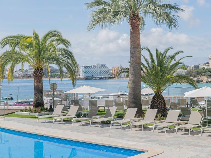 Espace piscine avec chaises longues et palmiers avec vue sur la mer et des bâtiments en arrière-plan.