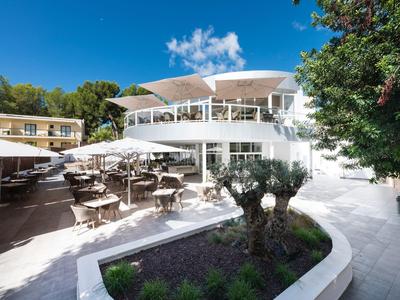 Modernes weißes Hotelgebäude mit Terrasse und vielen Tischen unter blauem Himmel.