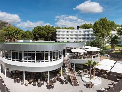 Modernes Hotel mit rundem Restaurant, Sonnenterrasse und umgeben von Bäumen bei blauem Himmel.
