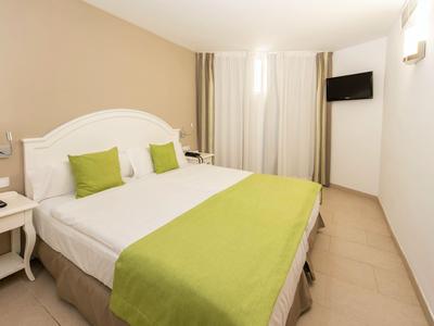 Hotelzimmer mit großem Doppelbett, grüner Bettdecke und Kissen sowie wandmontiertem Fernseher.