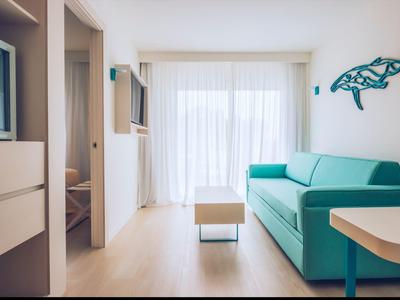 Helles Wohnzimmer mit türkisfarbenem Sofa, kleinem Tisch, heller Holzboden und großer Fensterfront.