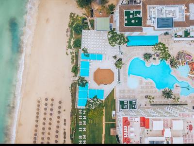 Luftaufnahme eines Strandes neben einem großen Poolbereich mit Liegen, Palmen und Sandstrand.
