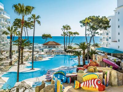 Großer Pool mit bunten Liegen, Palmen und Blick auf das blaue Meer bei sonnigem Wetter.