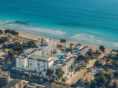 Luftaufnahme eines Hotels mit Pool am Sandstrand und blauem Meer, umgeben von Palmen und Gebäuden.
