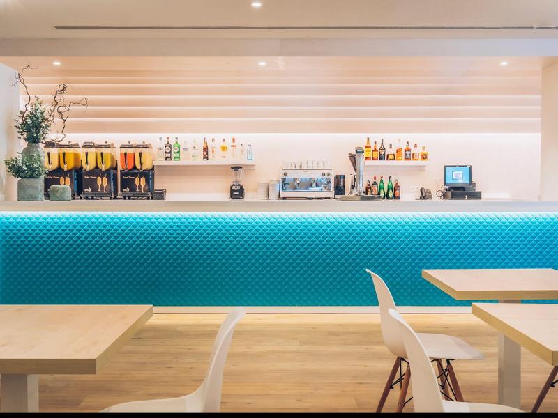 Lichte hotelbar met blauw verlichte bar en moderne houten tafels en stoelen.