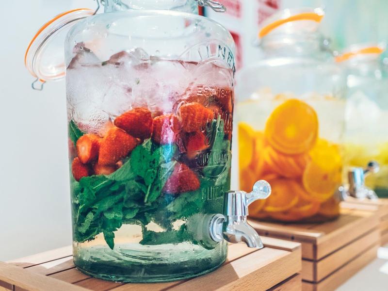 Drankdispenser met water, munt en aardbeien, andere met sinaasappels en citroenen op de achtergrond.