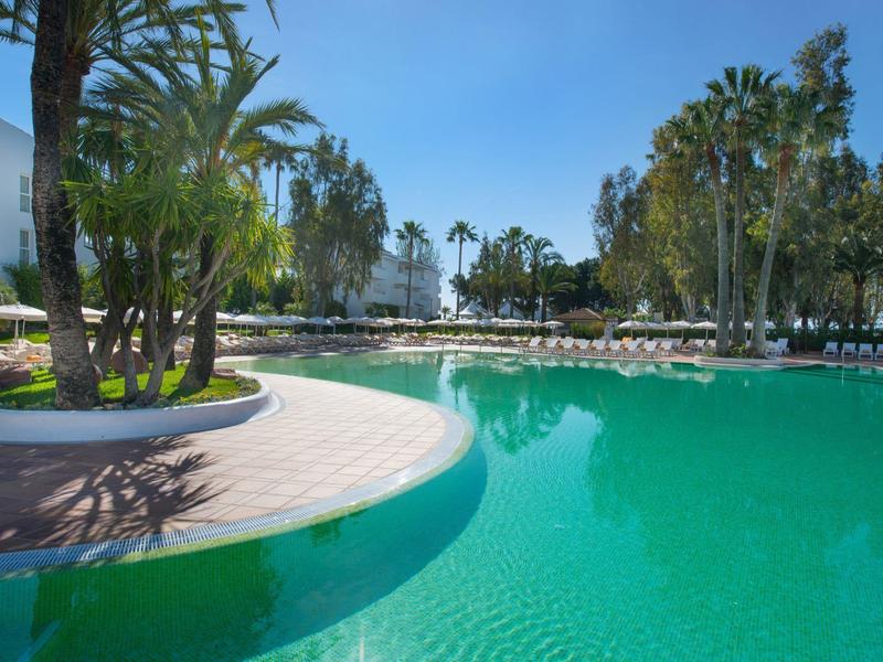 Großer Schwimmbadbereich mit türkisfarbenem Wasser und Palmen unter klarem Himmel.