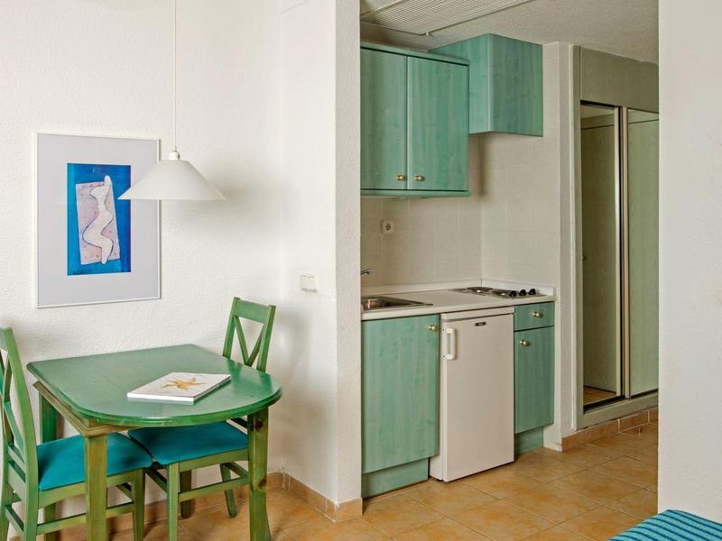 Kleine Küche mit grünen Schränken, grünem Essplatz und blau gestreiftem Bett im hellen Zimmer.