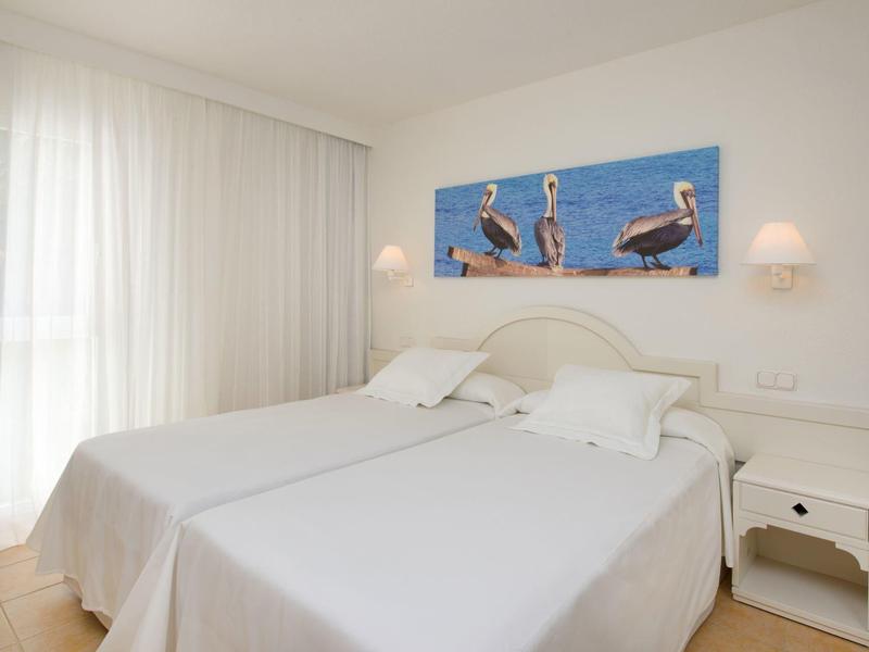 Helles Schlafzimmer mit zwei weißen Einzelbetten, weißem Nachttisch und Wandbild mit Pelikanen.