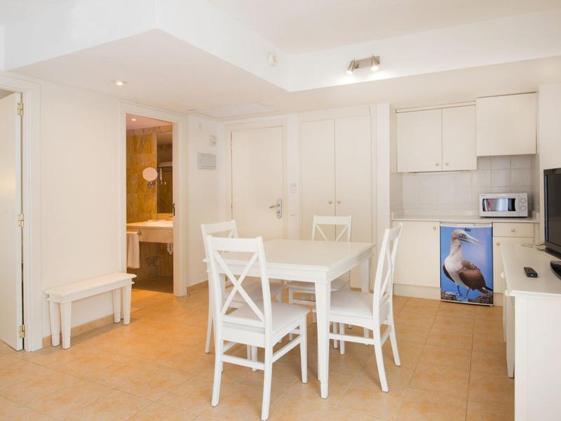 Heller Raum mit weißen Möbeln, Esstisch, vier Stühlen, kleinem Küchenschrank und Badezimmer im Hintergrund.