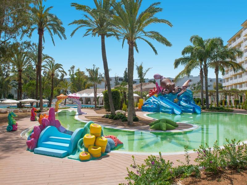 Kinderpool mit bunten Rutschen, Palmen und Hotelgebäude bei sonnigem Wetter.