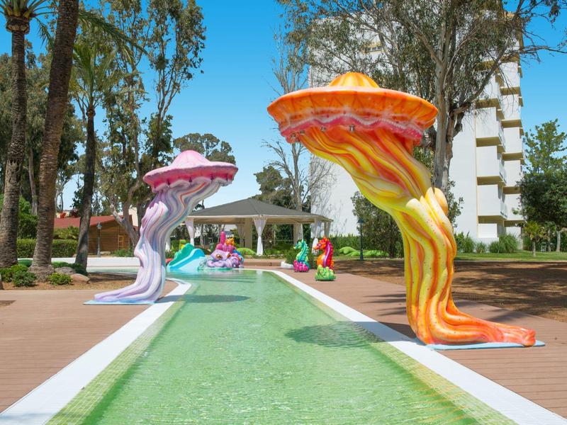 Bunte Riesenpilzskulpturen entlang eines grünen Wasserkanals im Park unter blauem Himmel.