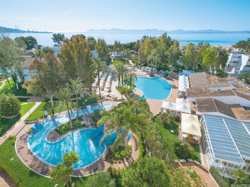 Hotelanlage mit zwei großen Pools, umgeben von Palmen, Wiesen und Blick auf das Meer im Hintergrund.
