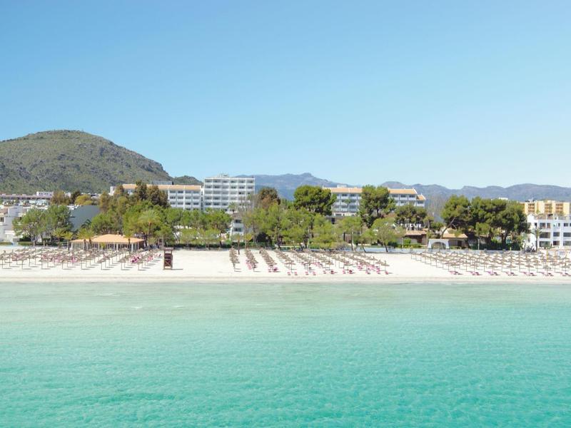 Klares türkisfarbenes Wasser, heller Sandstrand mit Liegestühlen, dahinter grüne Bäume und Hotels.