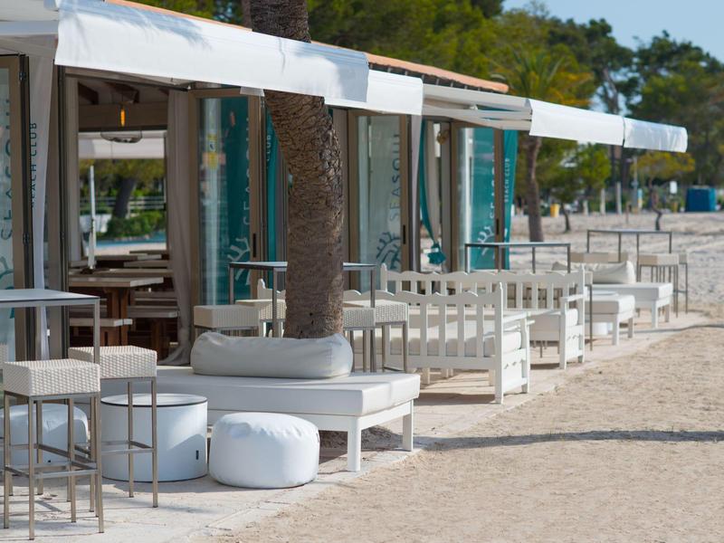 Strandbar mit weißen Sitzmöbeln und Sonnensegeln, umgeben von Sand und Bäumen im Hintergrund.