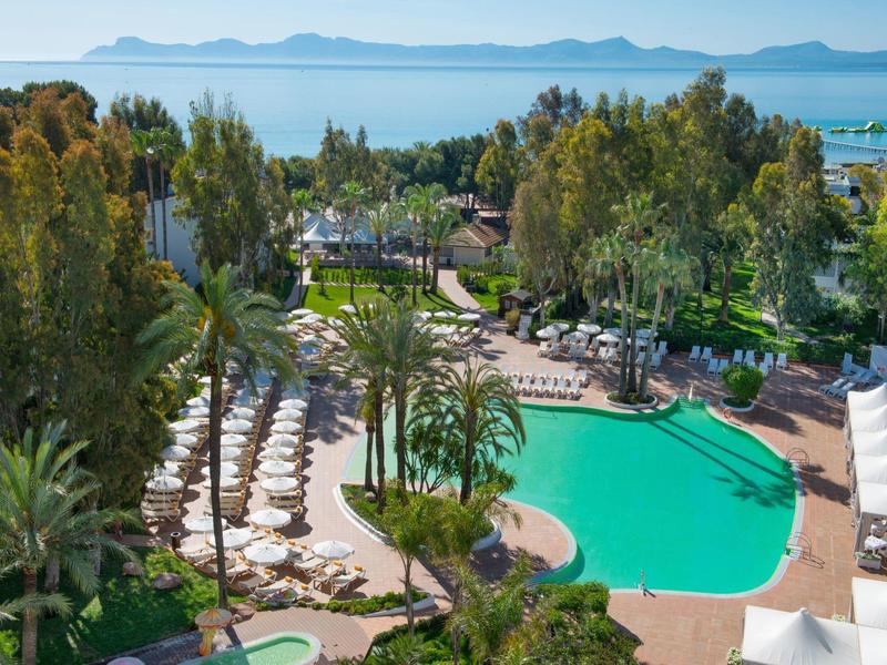 Großer Pool in tropischer Gartenanlage mit Palmen, Liegestühlen und Meer im Hintergrund.