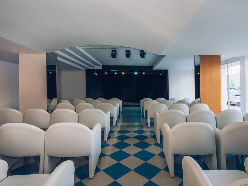 Leerer Veranstaltungsraum mit weißen Sesseln, blauer Bühne und kariertem Boden in Blau und Beige.