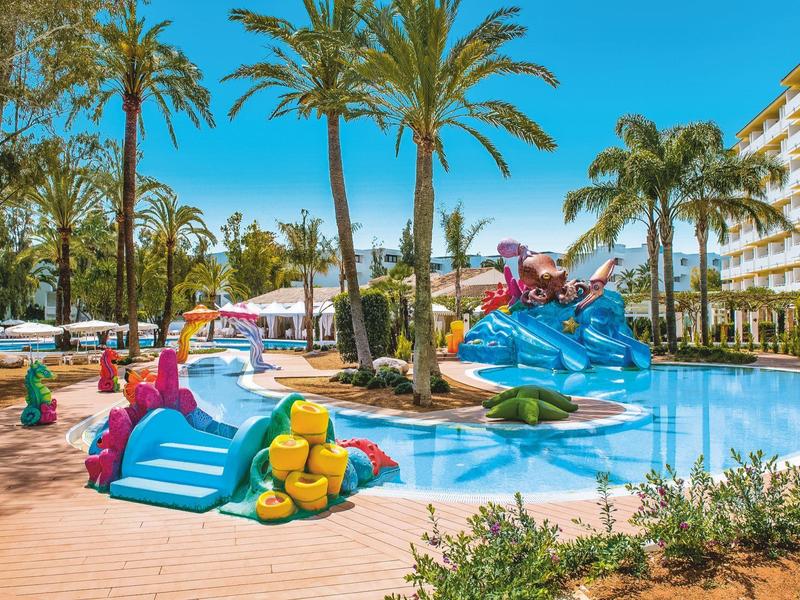 Bunter Wasserspielplatz mit Rutschen, Palmen und blauem Pool unter klarem Himmel.