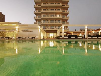 Una piscina iluminada frente a un edificio de hotel de varios pisos al anochecer.