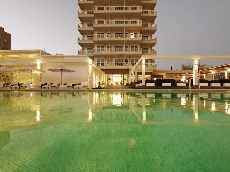Una piscina iluminada frente a un edificio de hotel de varios pisos al anochecer.