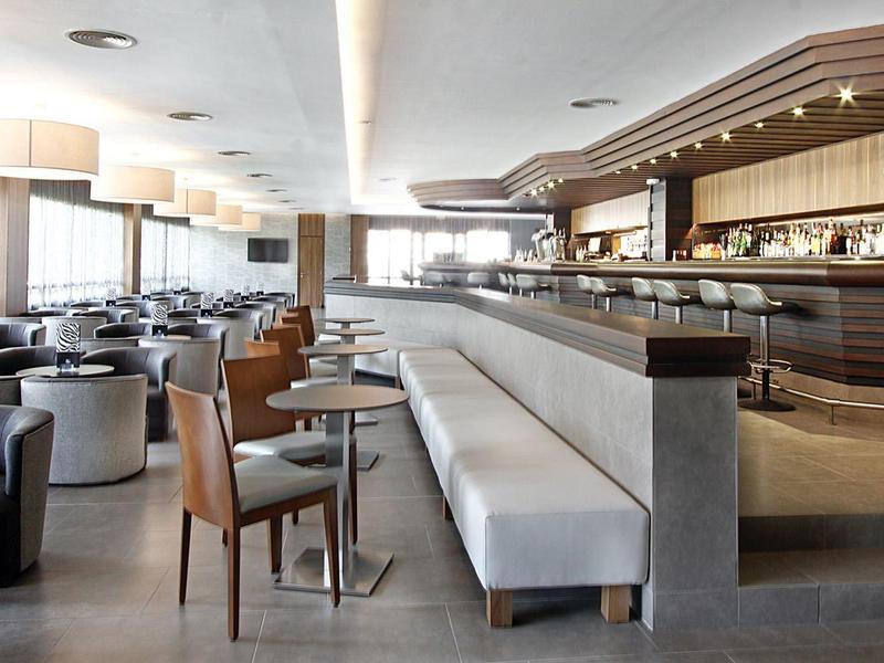 Área moderna de bar en hotel con asientos y grandes ventanas que permiten la luz natural.