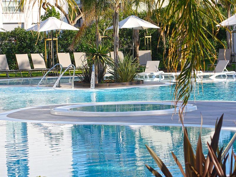Una piscina tranquila de hotel con sillas de descanso y sombrillas al fondo y palmeras al frente.