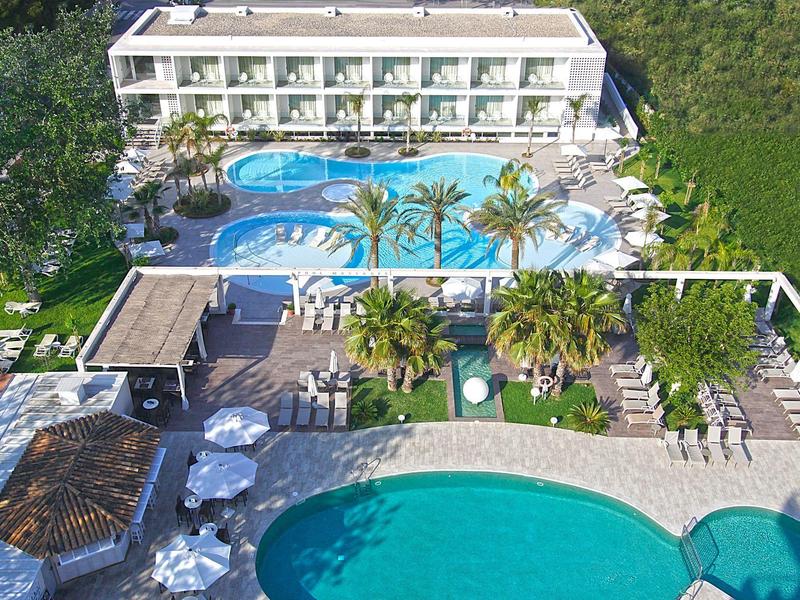 Hotel con dos áreas de piscina, rodeado de sombrillas y plantas verdes en un ambiente relajado.