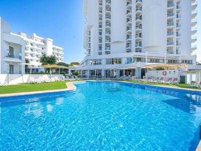 Duży zewnętrzny basen z czystą niebieską wodą przed białym, wielopiętrowym hotelem.