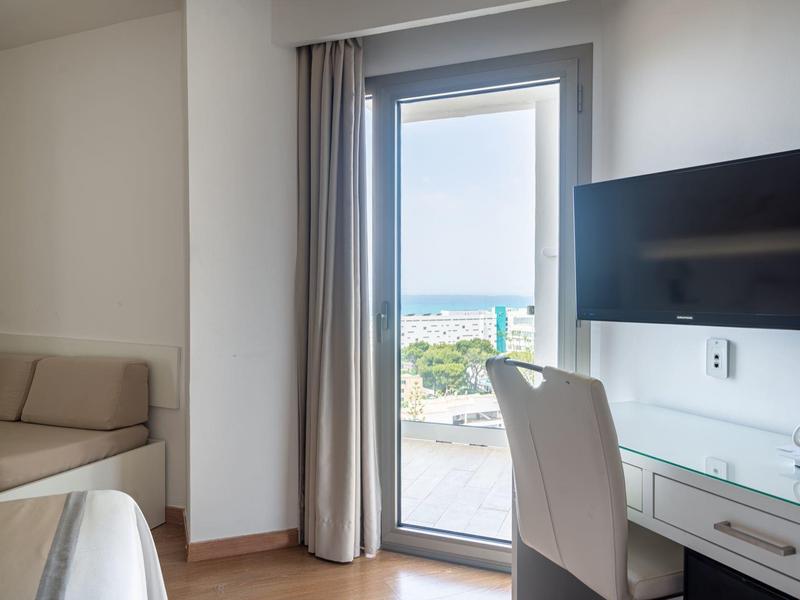 Jasny pokój hotelowy z biurkiem, krzesłem, telewizorem i dostępem do balkonu z widokiem na morze.