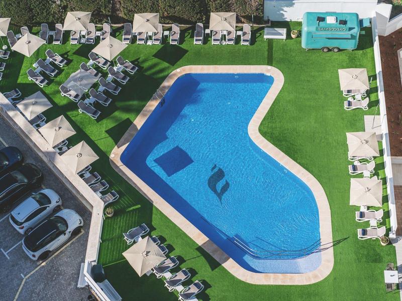 Widok z lotu ptaka na nieregularny basen z leżakami i parasolami na zielonej trawie obok miejsc parkingowych.
