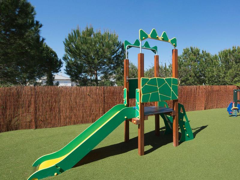 Zelenožlutá skluzavka na dětském hřišti s umělým trávníkem a stromy v pozadí.