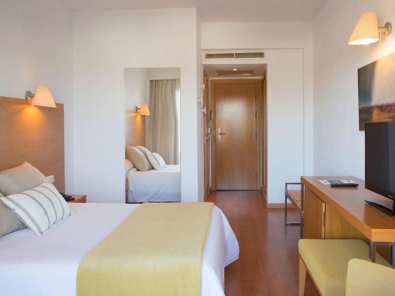 Světly hotelový pokoj s manželskou postelí, psacím stolem, židlí a nástěnným zrcadlem.