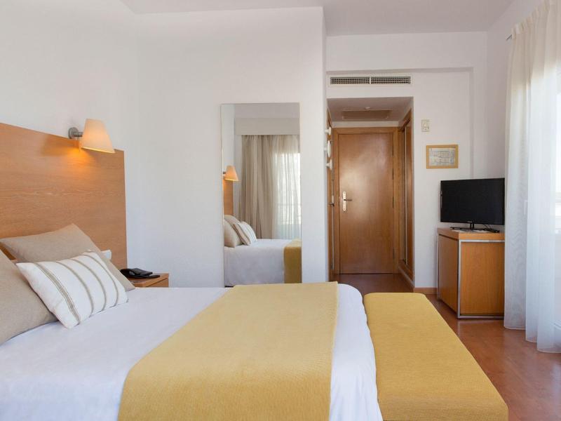 Světý hotelový pokoj s manželskou postelí, žlutou přikrývkou a televizí u dveří.