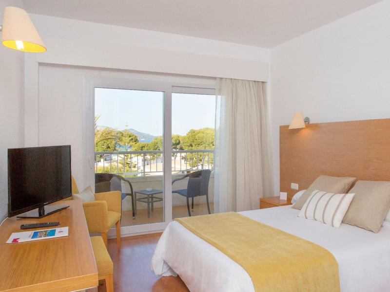 Světlý hotelový pokoj s manželskou postelí, psacím stolem, televizí a balkonem s výhledem na stromy.