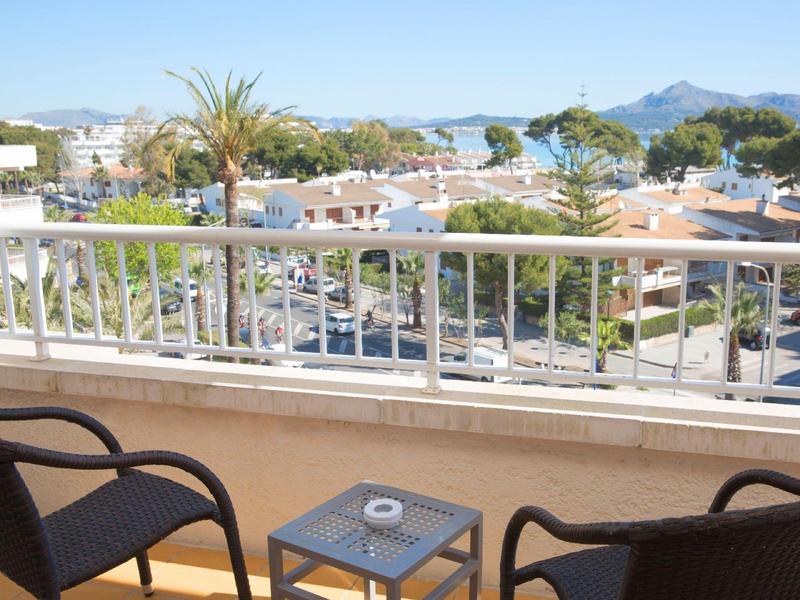 Balkon se dvěma židlemi a stolem s popelníkem, výhled na střechy a hory v pozadí.