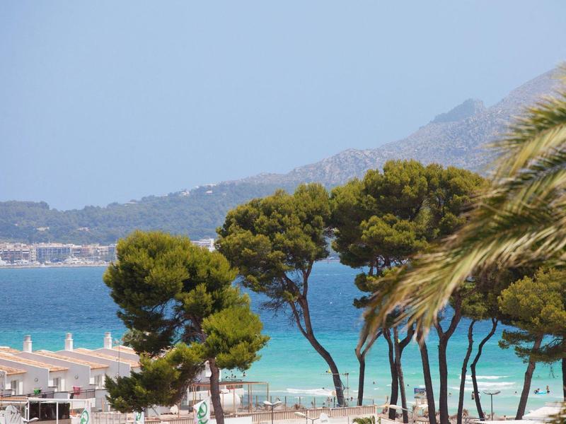 Pláž s čistou modrou vodou, stromy v popředí a hory v pozadí.