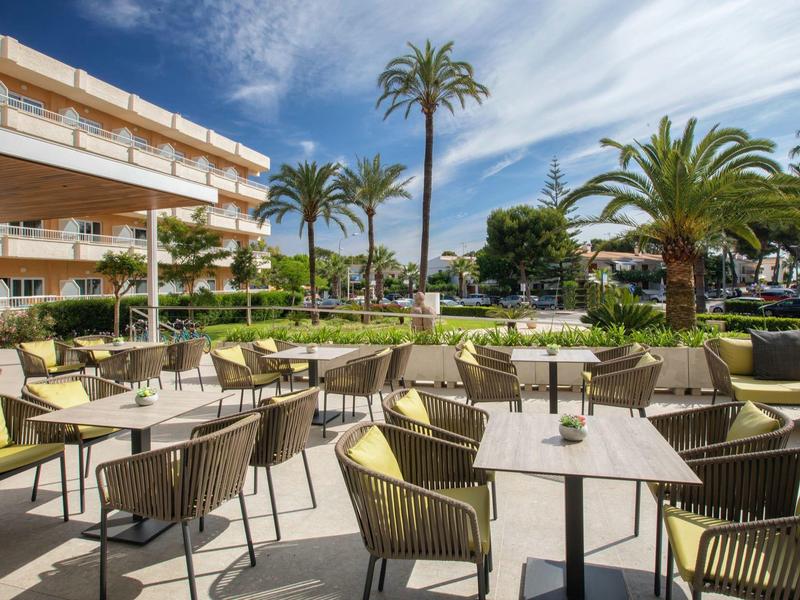 Venkovní posezení hotelu s stolem a zelenými židlemi pod palmami za slunečného dne.