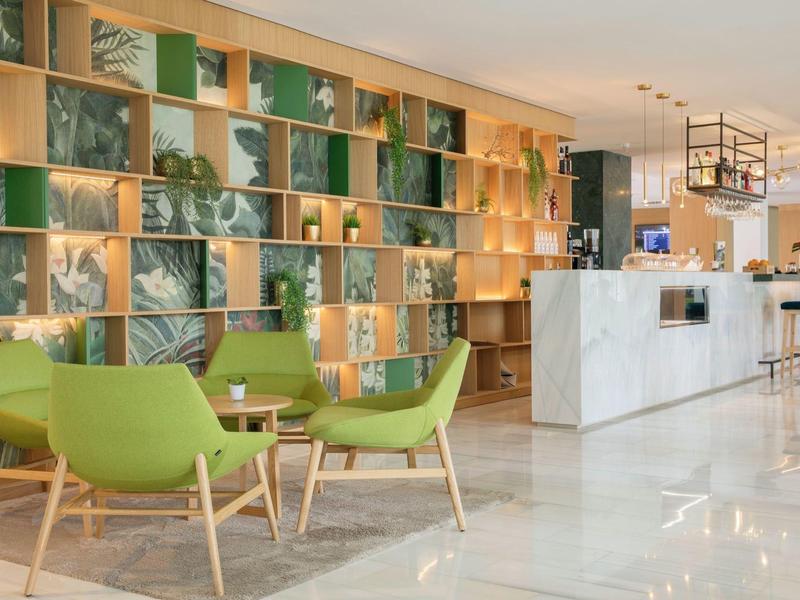 Světlý hotelový lobby s zelenými čalouněnými křesly, dřevěným nábytkem a bílou recepcí.