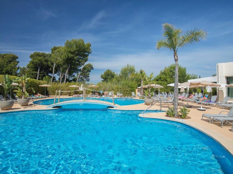 Velký venkovní bazén s lehátky, slunečníky a palmami pod jasnou oblohou.