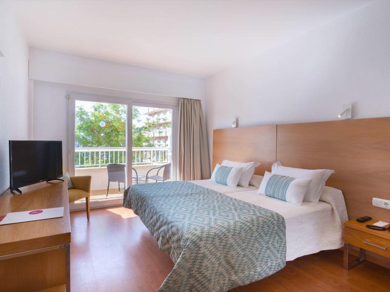 Světý hotelový pokoj s manželskou postelí, psacím stolem, TV a balkonem s výhledem do zeleně.