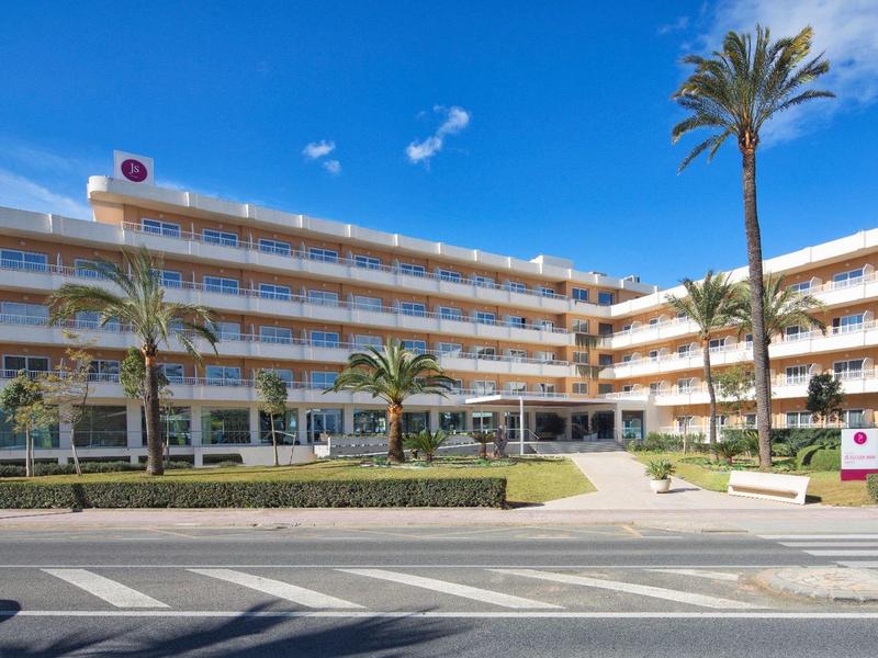 Moderní hotelová budova s několika patry, palmami a jasně modrou oblohou.