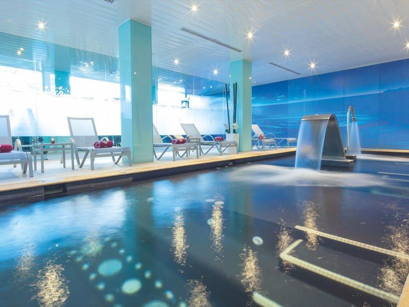 Moderní krytý bazén s vodní atrakcí a lehátky u velkého okna.