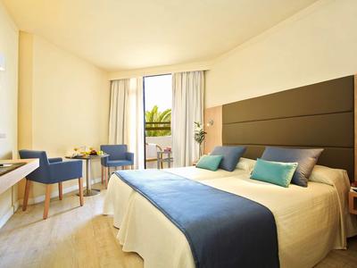 Lichte hotelkamer met tweepersoonsbed, blauwe accenten, stoelen en balkondeur.