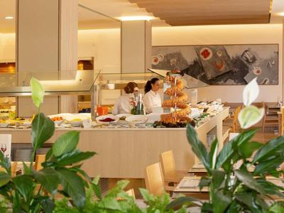 Moderne buffetruimte met koks die eten bereiden, houten stoelen en groene planten op de voorgrond.