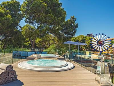 Terrazza in legno con jacuzzi, asciugamani, ombrellone, piscina e alberi sotto il cielo azzurro.